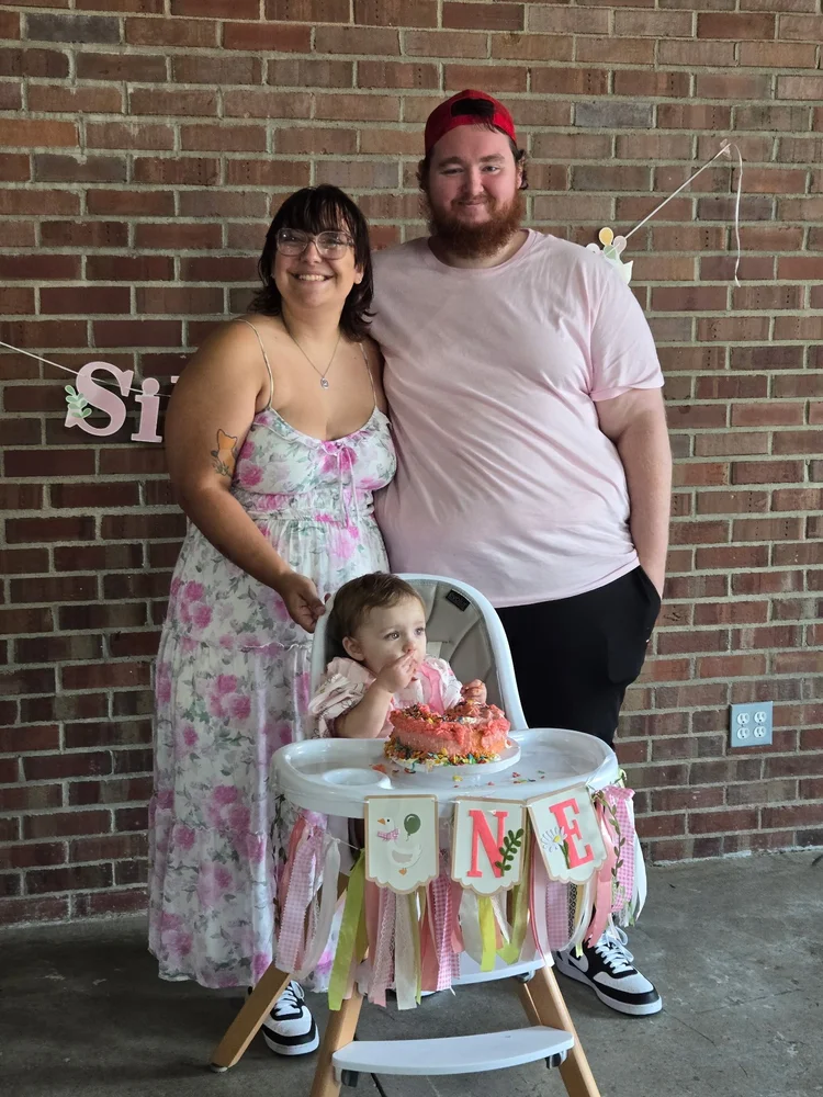 Two adults standing behind a child seated at a decorated birthday table in front of a brick wall.