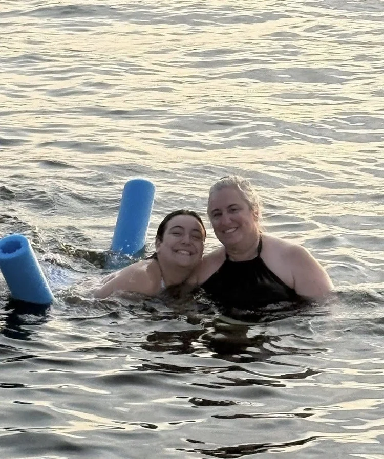 Two people smiling while swimming in open water with blue pool noodles.