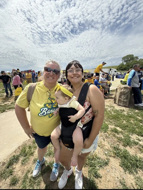 Two women standing outdoors with a child in a carrier, with people in the background.