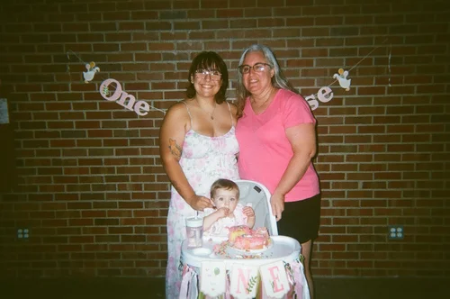 Two women standing behind a child seated at a decorated birthday table in front of a brick wall.