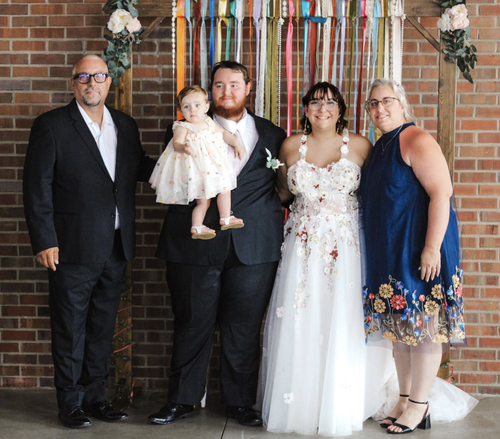 Five people posed at a wedding, including a child held by an adult, in front of a brick wall.
